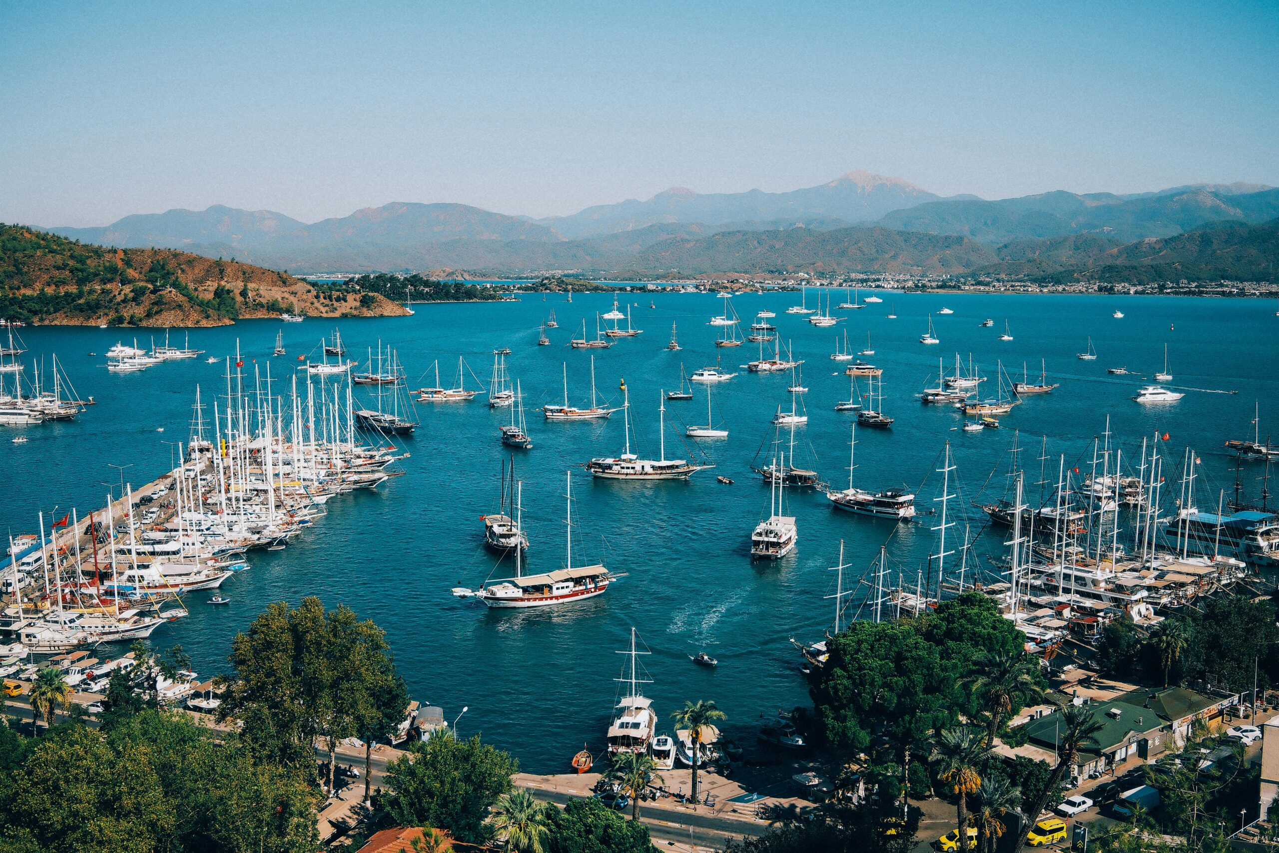 Explore the vibrant Fethiye Marina with dozens of colorful boats docked against a mountain backdrop.